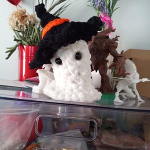 Puede incluir: Un fantasma blanco de ganchillo hecho a mano que lleva un sombrero de bruja negro con una banda naranja. El fantasma tiene ojos negros grandes y una textura esponjosa. El fondo incluye una decoración en forma de corazón rojo y varios arreglos florales.