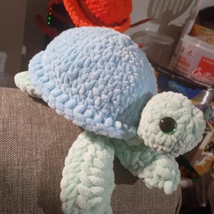 Puede incluir: Una tortuga de peluche hecha a mano en tonos azules y verdes. La tortuga tiene un caparazón azul claro, aletas verdes y una cabeza verde con ojos grandes y oscuros. El peluche está hecho de un material suave y texturizado.