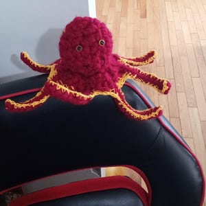 Puede incluir: Un juguete de pulpo de peluche hecho a mano en color rojo oscuro con ribetes amarillos en los tentáculos. El pulpo tiene dos ojos redondos y marrones y está sentado en una silla negra con detalles rojos. El juguete está hecho de un material suave y texturizado.