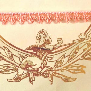 May include: Pink crocheted trim with a scalloped edge. The trim is against a background of a gold-colored design of a cherub.