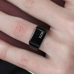 Op de afbeelding: Een zwarte rechthoekige ring met de letters "XZB" gegraveerd. De ring wordt om een vinger gedragen. De achtergrond omvat een bordeauxrood leren item en een zwart kledingstuk.