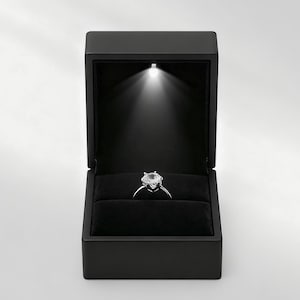 Könnte beinhalten: Eine offene, schwarze Schmuckschatulle mit einem Diamantring, der von einem Scheinwerfer beleuchtet wird. Der Ring liegt auf einem schwarzen Samt. Die Schatulle ist rechteckig mit einem Klappdeckel.