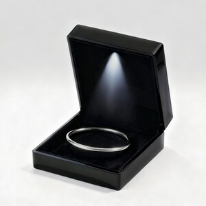 May include: A silver bangle bracelet rests inside a black jewelry box. The box is open, revealing a black velvet interior and a spotlight shining on the bracelet. The box has a glossy finish, and the background is white.