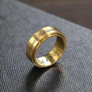 Op de afbeelding: Een goudkleurige ring met gegraveerde tekst. De ring heeft de naam "Damian Hellstrom" en de datum "8.4.2025". De ring ligt op een grijze, getextureerde achtergrond.