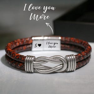 Puede incluir: Una pulsera de cuero marrón con un cierre de nudo plateado. La pulsera tiene un cierre magnético con el texto "I love you More" y la letra "S" con un símbolo de corazón.