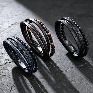 May include: Three multi-strand bracelets featuring braided leather and beaded accents. Each bracelet has a black magnetic clasp engraved with two hearts and the text LOVE FOREVER. The beads include matte black, tiger eye, and blue stone variations.