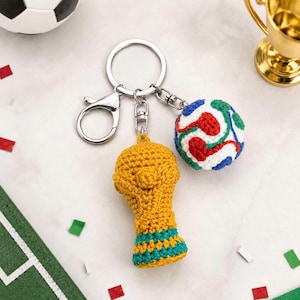 May include: A keychain featuring a gold-colored crochet trophy and a crochet soccer ball with red, green, blue, and white accents. The keychain also includes a silver clasp and key ring.