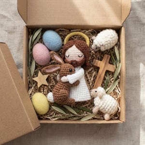 May include: A brown cardboard box filled with Easter-themed crochet items. Includes a Jesus doll, a brown bunny, two white sheep, a wooden cross, a wooden star, and three pastel-colored eggs. The box is lined with brown paper and greenery.