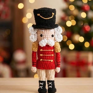 Könnte beinhalten: Handgefertigte Nussknacker-Puppe aus Häkelarbeit. Die Puppe hat einen schwarzen Zylinder, eine rote Jacke mit goldenen Knöpfen und schwarze Stiefel. Das Gesicht ist beige mit weißen Haaren und einem weißen Schnurrbart. Der Hintergrund ist verschwommen mit einem Weihnachtsbaum.
