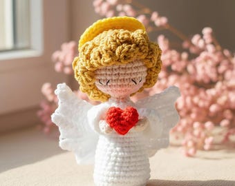 May include: A handmade crochet angel figurine. The angel has a yellow halo, curly blonde hair, and closed eyes. It wears a white dress and holds a red heart. White wings are attached to the back. The figurine is displayed on a neutral surface.