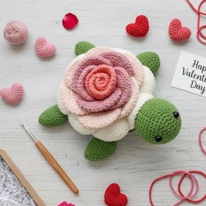 Könnte beinhalten: Eine handgefertigte Häkelschildkröte mit einem rosenförmigen Panzer in Rosa und Weiß, grünen Flossen und einem grünen Kopf. Kleine rote und rosa gehäkelte Herzen, ein Wollknäuel und eine Karte mit der Aufschrift "Happy Valentine's Day" sind verstreut.