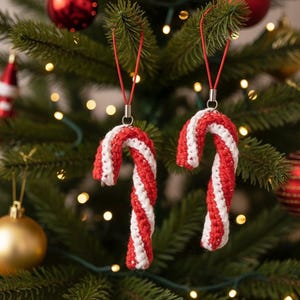 May include: Two red and white crocheted candy cane Christmas ornaments hanging from a green Christmas tree. The candy canes have a red and white striped pattern and are suspended by red strings. Other ornaments and lights are visible in the background.