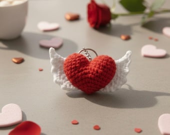 May include: A red crocheted heart with white wings attached to a silver keychain. The heart-shaped keychain is surrounded by small pink and red heart-shaped decorations, and a red rose.