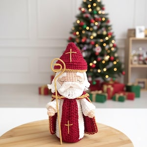 Può includere: Figura di San Nicola all'uncinetto fatta a mano, con cappello rosso e croce, barba bianca e bastone dorato. La figura indossa abiti rossi e bianchi. Albero di Natale e regali sullo sfondo.