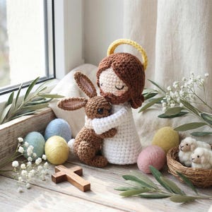 May include: A crocheted figure of Jesus with a brown beard and halo, hugging a brown bunny. Pastel-colored Easter eggs, a wooden cross, and two small white sheep are also present. The scene is set on a wooden surface with greenery.