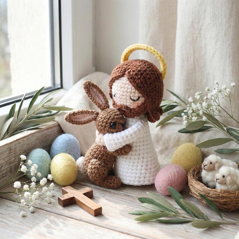 May include: A crocheted figure of Jesus with a brown beard and halo, hugging a brown bunny. Pastel-colored Easter eggs, a wooden cross, and two small white sheep are also present. The scene is set on a wooden surface with greenery.