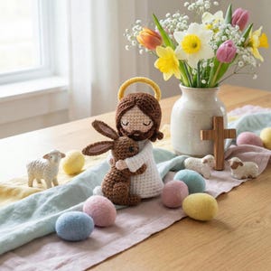 May include: A crocheted Jesus doll with a brown bunny, surrounded by pastel Easter eggs and ceramic lambs. A vase of spring flowers and a wooden cross are also on the table, creating a festive Easter scene.