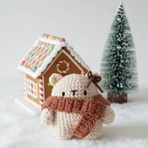 May include: A crocheted bear wearing a brown scarf and a small bow. A gingerbread house and a miniature Christmas tree are in the background. The bear is cream-colored and has small black eyes.