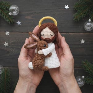 May include: A handmade crochet doll of a figure with brown hair, a yellow halo, and a white robe, hugging a brown bunny. The doll is held in two hands. The background includes Christmas ornaments and pine branches.