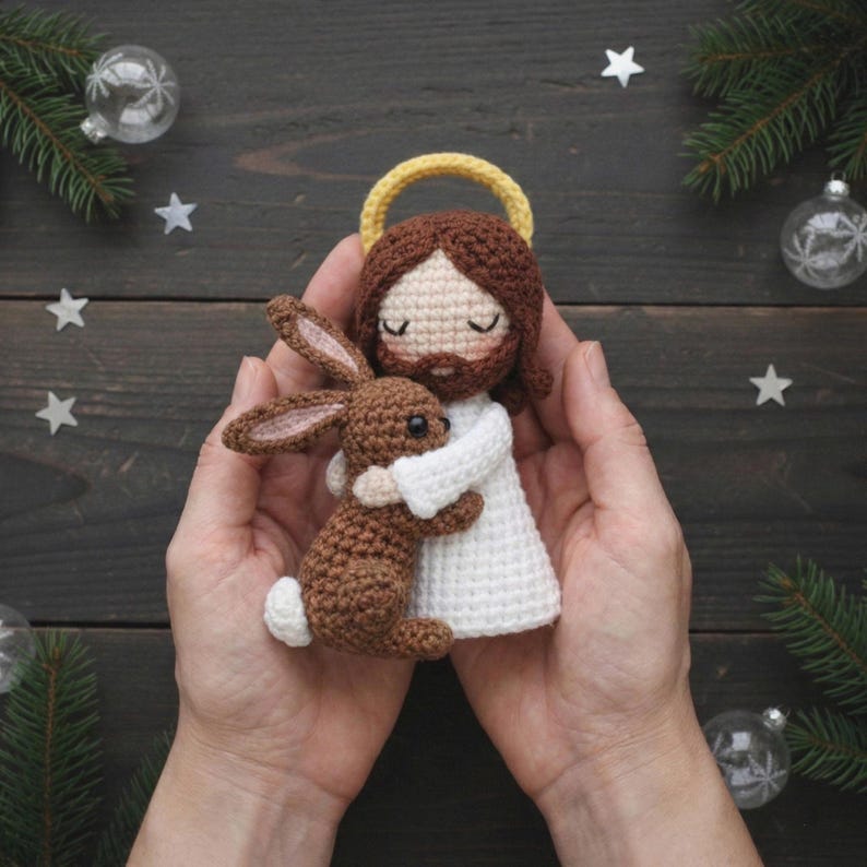 May include: A handmade crochet doll of a figure with brown hair, a yellow halo, and a white robe, hugging a brown bunny. The doll is held in two hands. The background includes Christmas ornaments and pine branches.