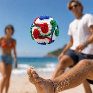 May include: A spherical, hand-crocheted ball featuring a white base with intricate, swirling patterns in vibrant red, blue, and green yarn. The textured, soft-looking ball is captured mid-air, showcasing its detailed stitch work and colorful design.