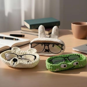 May include: Three handmade crochet eyeglass holders shaped like a bear, a bunny, and a crocodile. The bear holder is off-white with a black nose. The bunny holder is off-white with tall ears. The crocodile holder is green with white eyes.