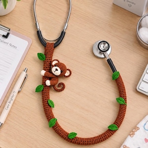 May include: A medical stethoscope featuring a brown crocheted cover on the tubing, designed to resemble a tree branch with small green crocheted leaves attached. A small, brown and cream-colored crocheted monkey is attached to the tubing.