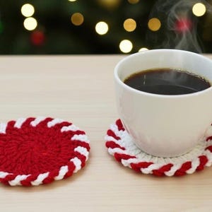 Op de afbeelding: Een witte keramische mok gevuld met donkere koffie staat op een rood-witte gehaakte onderzetter. Een andere onderzetter staat naast de mok. De onderzetters hebben een zuurstokontwerp. Stoom stijgt op uit de koffie.