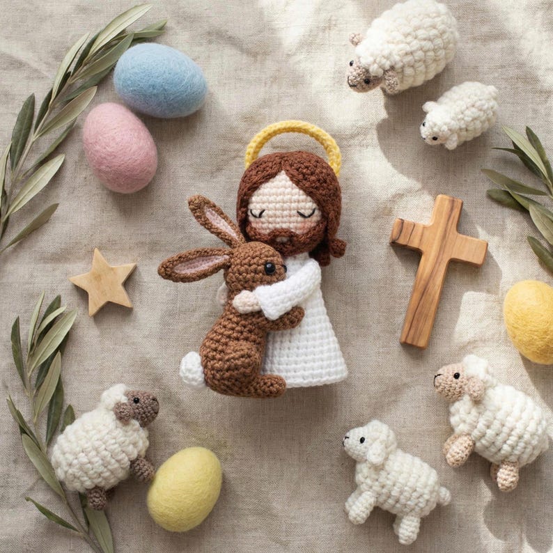May include: A collection of handmade Easter decorations. The centerpiece is a crocheted figure of Jesus embracing a brown bunny. Surrounding them are crocheted sheep, wooden cross, star, and felt eggs in pastel colors. Olive branches add a natural touch.