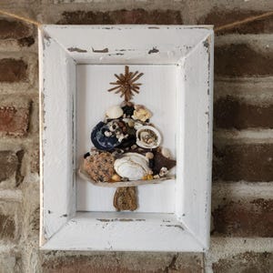 Può includere: Un'opera d'arte incorniciata con un design ad albero di Natale realizzato con varie conchiglie e piccole pietre. L'albero è posto su uno sfondo bianco all'interno di una cornice di legno bianco anticato. L'opera è appesa a un muro di mattoni. Dimensioni: circa 20 cm x 15 cm.