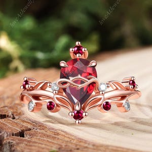 May include: Rose gold ring with a heart-shaped, deep red gemstone at the centre, complemented by smaller red and clear stones. The ring has an intricate design with a crown-like top, set against a natural wood background.