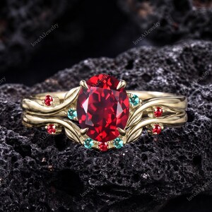 Puede incluir: Un anillo de oro con una gran piedra central de color rojo intenso y corte ovalado, acentuado por piedras redondas más pequeñas en rojo y turquesa engastadas en una banda con motivos metálicos de hojas que rodean el centro.