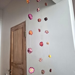May include: A decorative floral garland featuring dried flowers in shades of pink, orange, and burgundy, suspended from the ceiling. The flowers are strung on thin, nearly invisible threads, creating a floating effect. A wooden door is visible in the background.