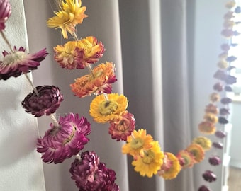 May include: A garland of dried flowers in shades of yellow, orange, and purple hangs against a neutral background. The flowers are strung together, creating a decorative chain. The flowers are in full bloom.