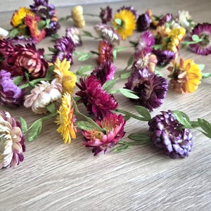 Op de afbeelding: Een verzameling gedroogde bloemen in verschillende kleuren, waaronder geel, paars en bordeauxrood, met groene bladerranken. De bloemen zijn gerangschikt op een lichtgekleurd houten oppervlak, wat een natuurlijke en rustieke uitstraling creëert.