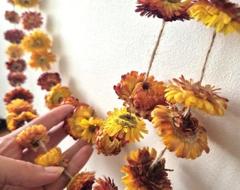May include: Dried flower garland featuring yellow, orange, and brown blooms strung on twine. The flowers are arranged in a decorative pattern, with some hanging individually. The garland is a natural, rustic home decor item.