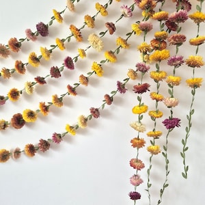 May include: Multiple vertical garlands featuring dried strawflowers in shades of yellow, orange, pink, and deep red. Each flower is attached to a thin green stem with small, pointed leaves, creating a cascading floral display of natural colors.