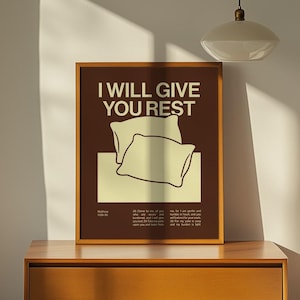 以下が含まれることがあります： 「I WILL GIVE YOU REST」のテキストと2つの枕のイラストが描かれた額入りのポスター。ポスターは茶色で、クリーム色のテキストとイラストが施されています。木製のナイトスタンドに飾られています。