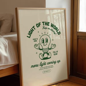 Può includere: Una stampa incorniciata con uno sfondo beige chiaro e un'illustrazione verde di una lampadina. Il testo recita "LIGHT OF THE WORLD" e "more light coming up". La cornice è di colore legno chiaro e la stampa è appoggiata a un muro.