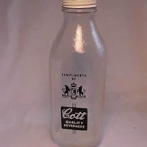 May include: A clear glass milk bottle with a white lid and a black and white label that reads "Compliments of Cott Quality Beverages".