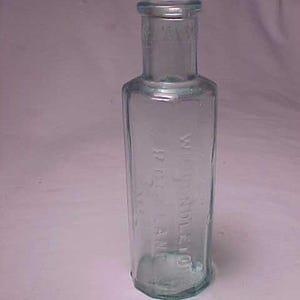 May include: A clear, rectangular glass bottle with a rounded base and a narrow neck. The bottle has embossed text on the side, and it is displayed against a white background. The bottle is empty and appears to be an antique.