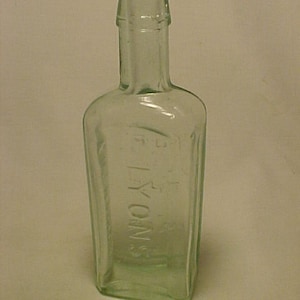 May include: A clear glass bottle with a rounded top and a square body. The bottle has the word "LYONS" embossed on the front.