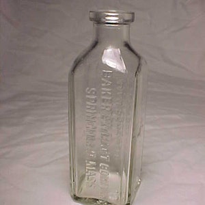 May include: A clear glass bottle with a square shape and a textured surface. The bottle is embossed with the text "Baker Extract Co. Springfield, Mass."