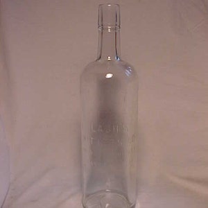 May include: A clear glass bottle with a long neck and a cylindrical body. The bottle has a slightly rounded base and a narrow opening. The text "LASH'S BITTER S.O." is embossed on the bottle. The bottle is empty and appears to be vintage.