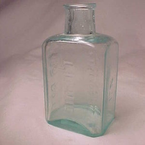 May include: A clear glass bottle with a square body and a rounded neck. The bottle has a textured surface and is slightly tinted green.