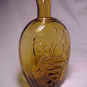 May include: A brown glass bottle with a rounded body and a short neck. The bottle has a raised design of a grapevine with grapes on the front.