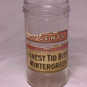 May include: A clear glass jar with a screw-on lid. The jar has two labels with text that reads "10 CENTS" and "FINEST TID BITS WINTERGREEN". The jar is cylindrical and appears to be vintage.