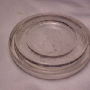 May include: A clear glass jar lid with a raised rim and a smooth, flat surface. The lid has a slightly frosted appearance.
