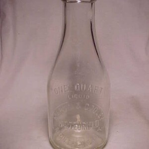 May include: A clear glass milk bottle with a rounded bottom and a narrow neck. The bottle is labeled "ONE QUART LIQUID" and "PASTEURIZED".