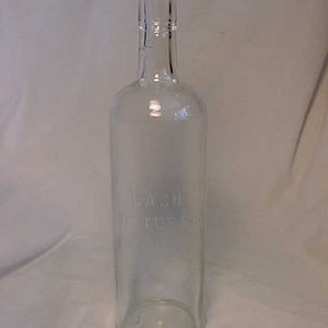 May include: A clear glass bottle with a textured surface. The bottle has the text "LASH" and "MILLER" etched into the glass.